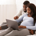 Young pregnant afro couple relaxing at home, using laptop, watching educational content for parents, free space